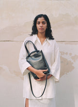 Load image into Gallery viewer, Pazar I Pazar Tote Nano Black I black I front view I showing iconic SAGAN closing system and strap detail I a model is wearing the bag in her hands in front of a public building