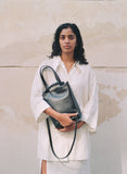 Load image into Gallery viewer, Pazar I Pazar Tote Nano Black I black I front view I showing iconic SAGAN closing system and strap detail I a model is wearing the bag in her hands in front of a public building